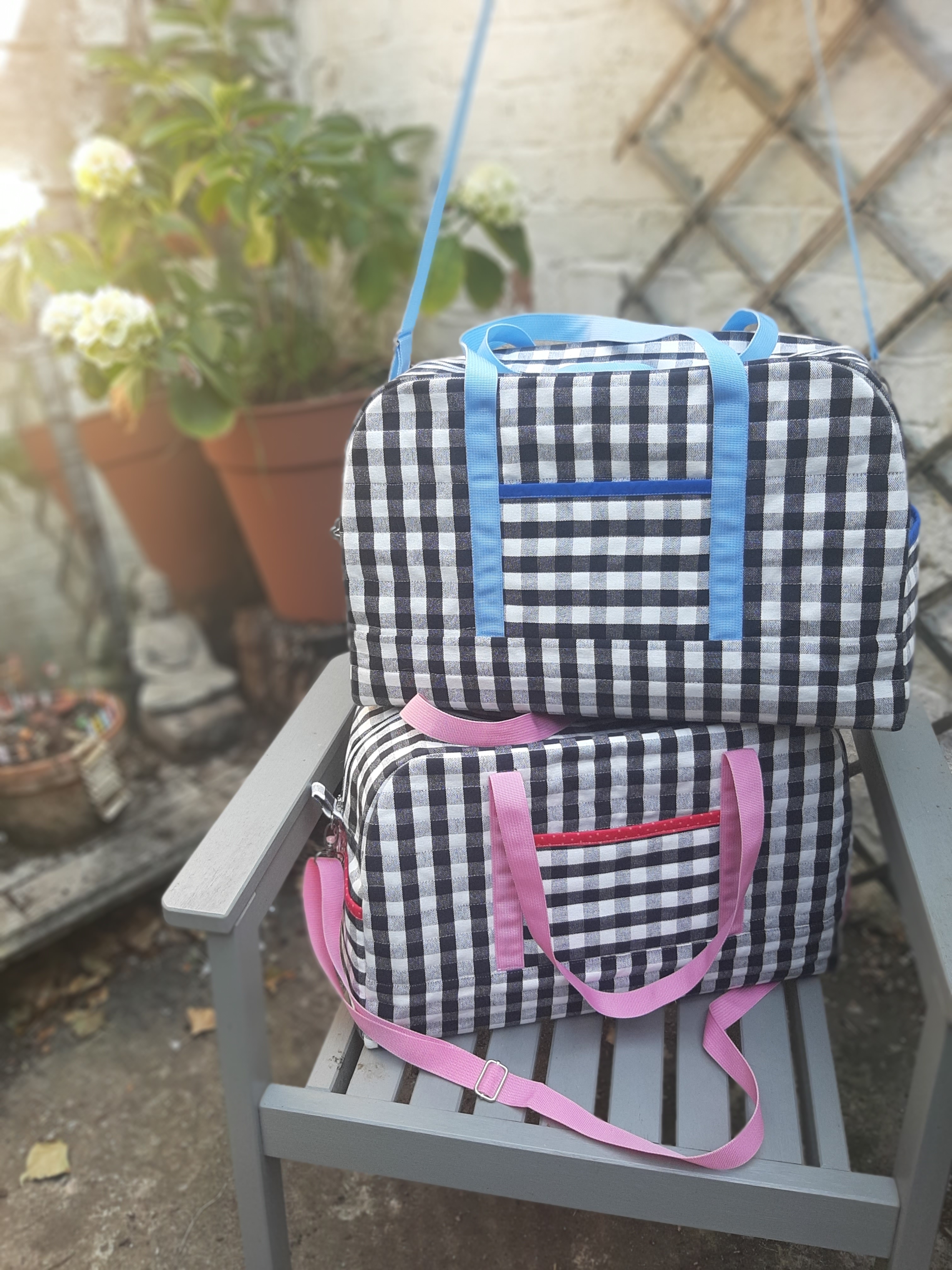 Outdoor photo of two similar luggage bags, one on top of the other. Both black and white gingham fabric, one with blue and one with pink as the accent colour.