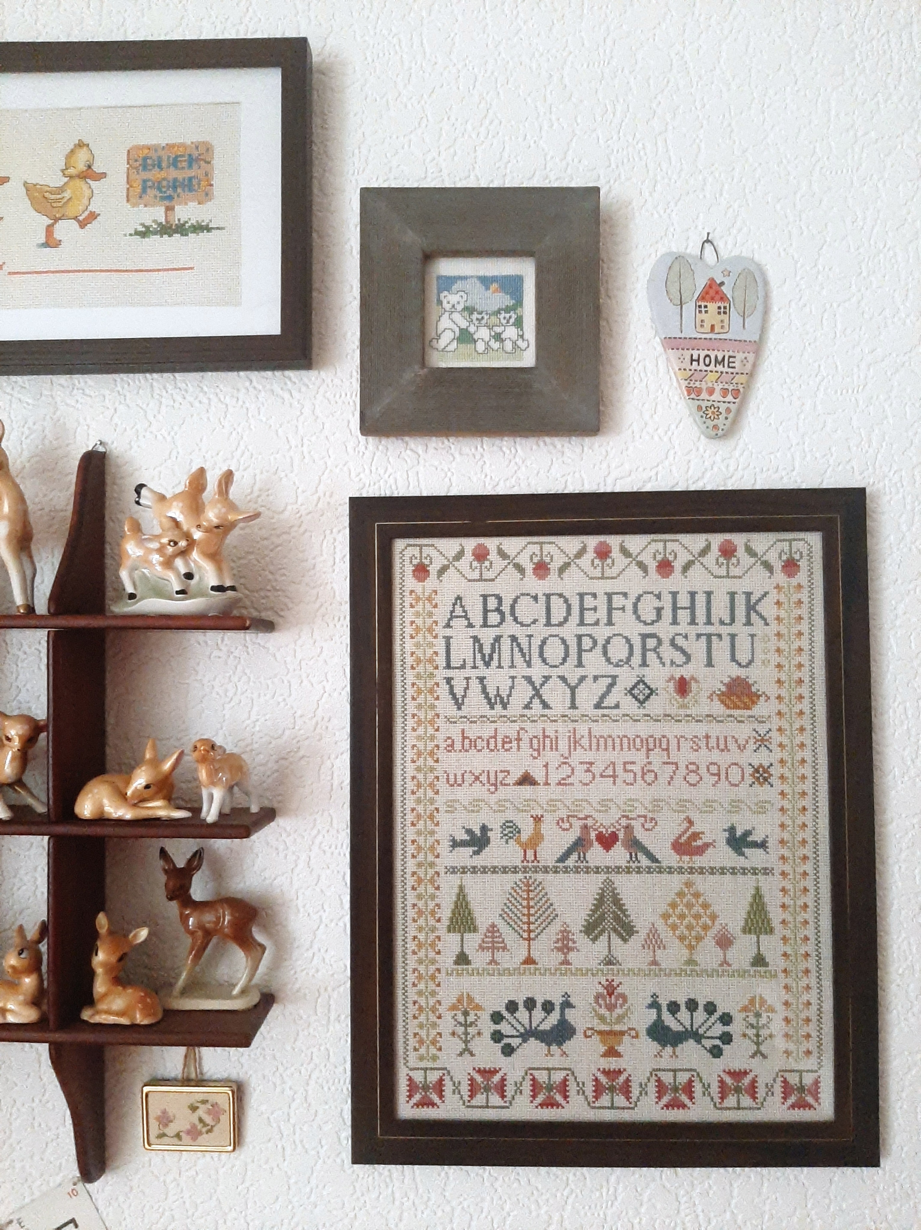 Assortment of cross-stitch pictures on a wall with a curio shelf filled with ceramic deer